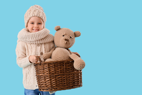 Cute little girl in winter clothes holding basket with toy bear on blue background