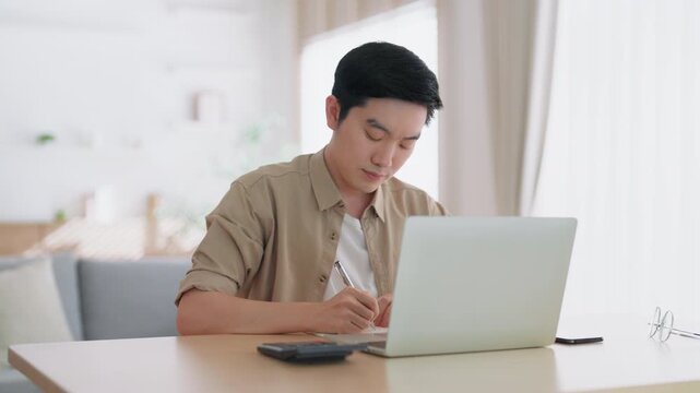 Happy young Asian man working on laptop in living room. Freelance worker male sitting at desk writing notes while watching webinar, studying online