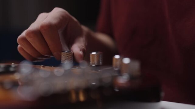 Close-up video showing musician hand turning chrome control knob on body of electric guitar. Scene has warm, focused lighting, creating moody and intimate atmosphere, closeup, slow motion.