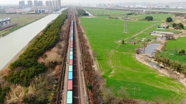 Aerial drone view of a long intermodal freight train with colorful shipping containers traveling along railway tracks beside a river and green farmland