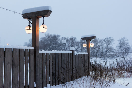 Glowing street lamps mounted on rustic wooden posts, topped with fresh white snow along a vintage picket fence during a calm and freezing winter evening in the countryside