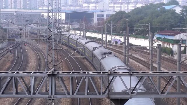 Red electric locomotive pulling a long passenger train along curved railway tracks, captured from an elevated metal bridge with urban buildings in the background