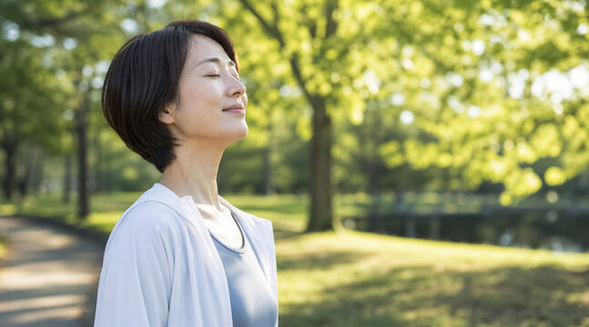 新緑の木漏れ日の中で目を閉じ深呼吸をして心を整えるミドル女性