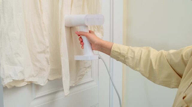 Woman using handheld garment steamer on a shirt hanging on a door. Household chore activity for wrinkle removal. Efficient garment care and laundry maintenance at home appliance concept.