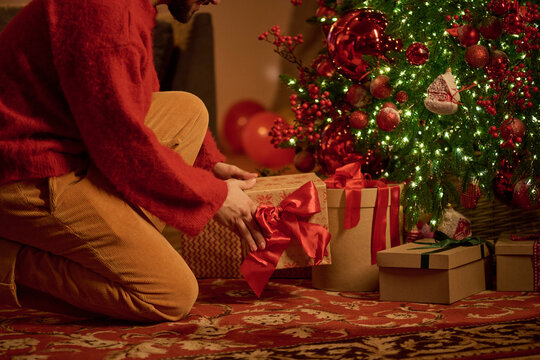 Young adult man placing wrapped gift box under decorated Christmas tree. Holiday celebration visual suitable for winter seasonal advertising campaigns