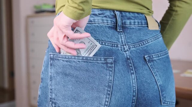 Woman tucks folded US dollar cash into rear pocket of jeans, guiding bills beneath stitched edge with one hand. Denim pocket holds money securely as she finishes putting banknotes away indoors