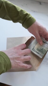 Woman puts stack of US dollar banknotes inside open kraft envelope on marble surface, arranging cash before folding paper flap over it for secure storage, delivery, or personal finance organization