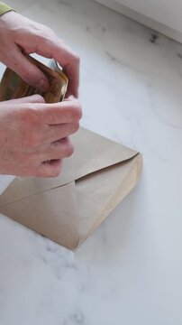 Hands put euro banknotes into open paper envelope, with woman arranging cash on pale marble counter and closing packet, placing gathered bills inside for household storage or safekeeping