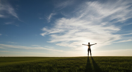 Obraz premium Silhouette of a person with arms outstretched in a vast green field under a beautiful blue sky with wispy clouds at sunset