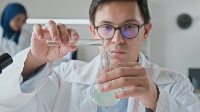 Asian young adult male scientist pouring chemical liquid from test tube into glass flask in modern laboratory. Footage suitable for pharmaceutical research and biotechnology marketing