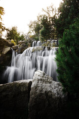 Scenic waterfall cascades over rugged rocks among lush green trees in soft sunlight, creating a tranquil landscape associated with nature, relaxation and fresh water. © Инна Мадеева
