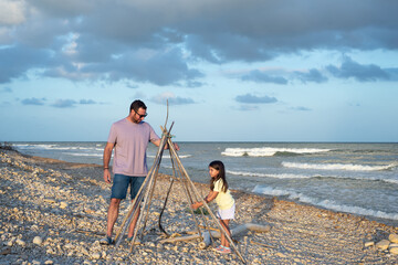 Father and young daughter building a teepee hut with natural canes on a pebble beach at sunset. © Esther Pueyo