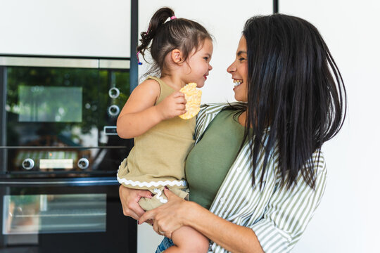 Happy mother holds her daughter eating a snack in the kitchen