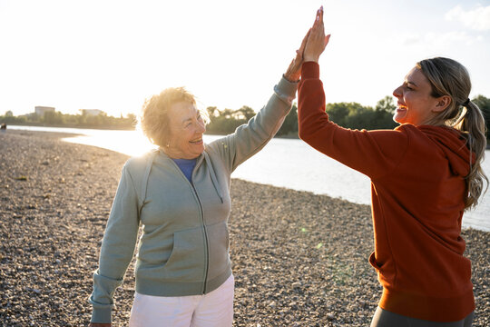 Senior woman and caregiver high five outdoors by the river at sunset
