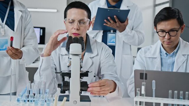 Middle aged Caucasian woman scientist using microscope and discussing clinical results with diverse research team. Suitable for pharmaceutical advertising, clinical trial report or biotech video