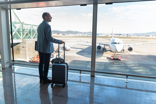 Businessman with suitcase looks at aircraft from airport terminal
