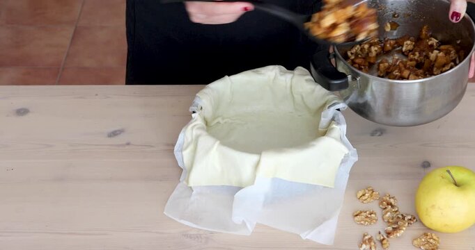 Cooked apple pieces flavored with cinnamon are transferred from a small saucepan into a puff pastry crust to prepare a homemade apple pie.