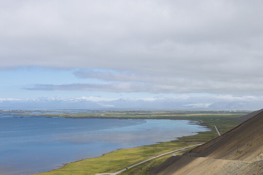 Elevated view from Almannaskard across Hornafjordur coastal water, green lowlands and a winding road toward distant snow covered mountains beneath layered clouds in southeast Iceland.
