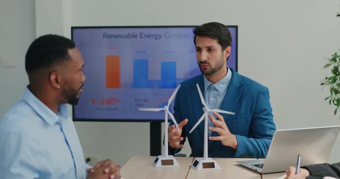 Portrait of Hispanic manager discusses renewable energy strategy near spinning turbine models with African American guy. Manager gestures and explains details