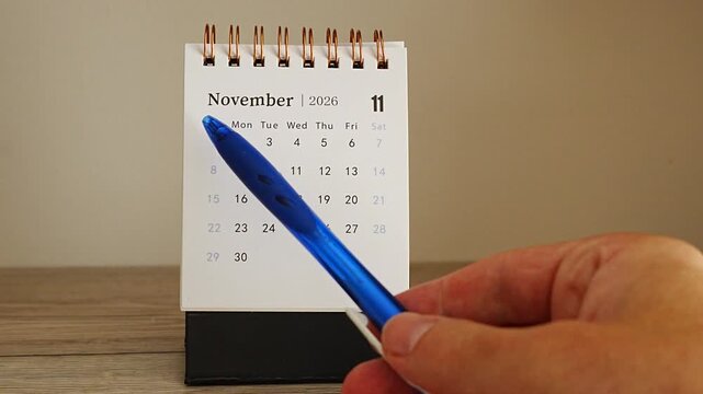 Hand holding a blue pen points to a specific day on a desk calendar page for November 2026, marking an important date on the spiral bound planner, concept of scheduling appointments and meeting deadl