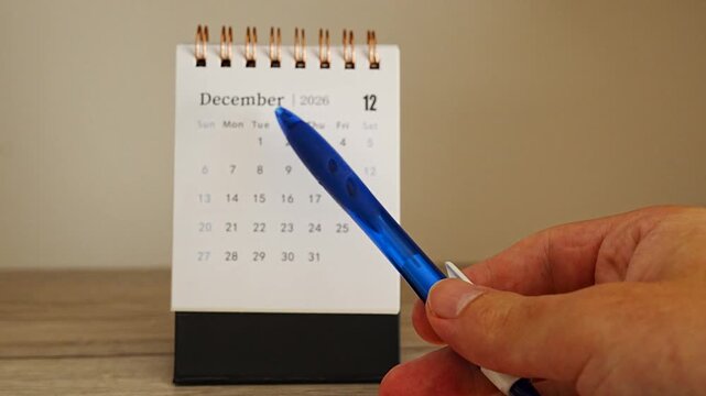 Pen tip pointing to the first week of December 2026 on a white desk calendar, hand entering from the right on a wooden desk against a warm beige wall, concept of start of the month and new tasks ahea