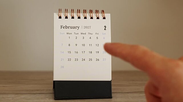 Finger approaching from the right edge toward a small white desk calendar showing February 2027, centered on a wooden table with plain beige wall and empty space around, concept of minimal planning a