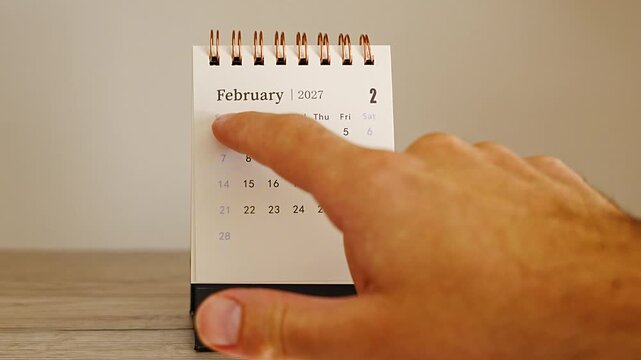 Index finger pointing at dates on a white desk calendar showing February 2027, spiral binding and wooden table against a soft beige background, concept of short month, Valentines Day planning and win