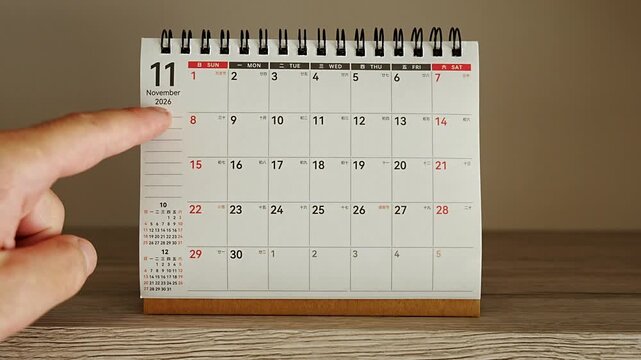 Hand tracing weekly rows on a wide monthly grid desk calendar for November 2026, red and black numbers with mini calendars on the side, concept of weekly planning, workload overview and organized rou