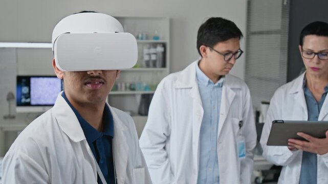 Young adult Indian scientist using VR headset with controller in laboratory beside multiethnic colleagues. Suitable for digital medical training and biotechnology innovation campaigns