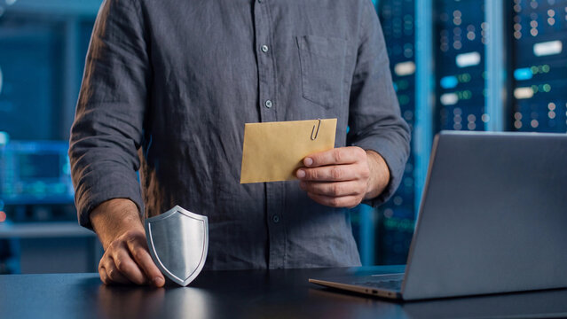 Email attachment security and inbox protection concept, man holding envelope with paperclip and metal shield near laptop in server room for phishing and malware defense