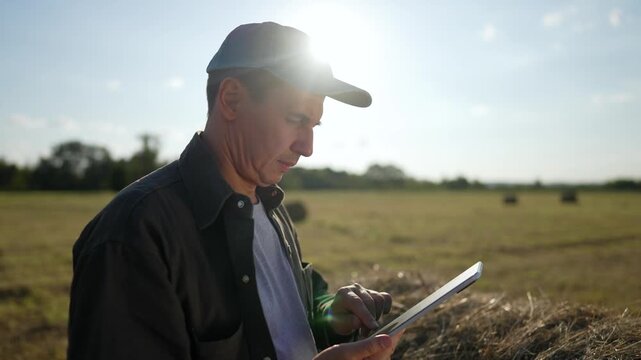 Happy male farmer smiles using tablet in hay field at sunset. Man farmer works on tablet with smile. Agricultural worker reviews data happily. Man smiles using tablet near hay bale outdoors.