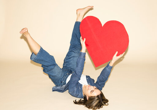 Barefoot woman in denim holding a large red paper heart up in the air