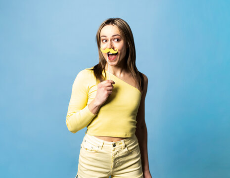 Woman holding a yellow paper mustache on a stick against a blue background