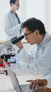 Vertical video of Asian male scientist looking through microscope with colleague typing research data on laptop nearby. Perfect for pharmaceutical studies and biotechnology advertising