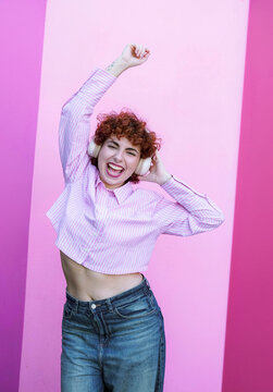 Smiling redhead listening to music and dancing against pink wall