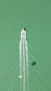 Top down aerial shot of speed motorboat towing parasail activity leaving white foam wake on turquoise sea surface with parachute shadow on water