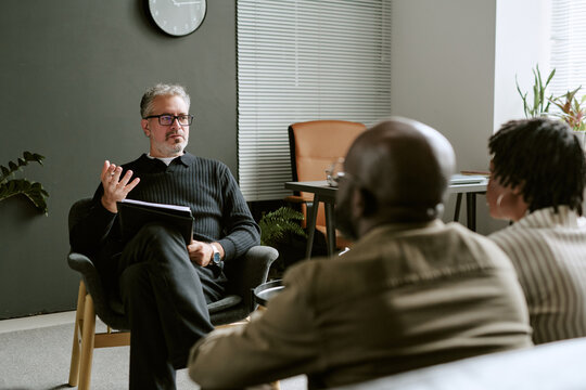 Middle aged Caucasian male psychologist gesturing during therapy consultation with African American couple. Image useful for mental wellbeing campaigns and family counseling services