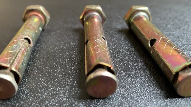 Real-time camera pan from left to right showing three yellow zinc plated sleeve anchor expansion bolts arranged vertically on a dark textured background. macro close up 4K video.