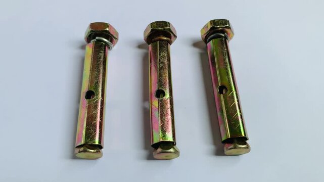 Real-time camera pan from left to right showing three yellow zinc plated sleeve anchor expansion bolts arranged on a clean white surface. High angle macro close up 4K video for masonry and hardware.