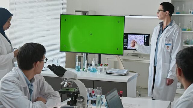 Middle aged Caucasian female scientist presenting research to multiethnic team using green screen monitor in laboratory. Footage suitable for pharmaceutical and biotechnology advertising