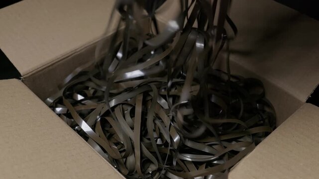 Unspooled magnetic audio tape ribbons pile loosely into an open cardboard box in close-up. The chaotic unraveling evokes vintage obsolescence, making it ideal for retro music documentaries.