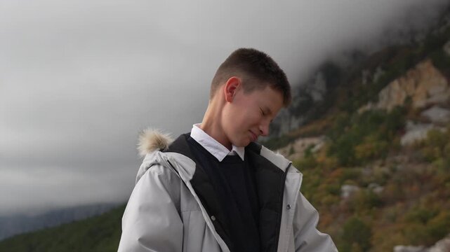 Boy mountain travel, young man smiles and laughs while wearing a warm winter jacket on a cloudy day overlooking scenic terrain.