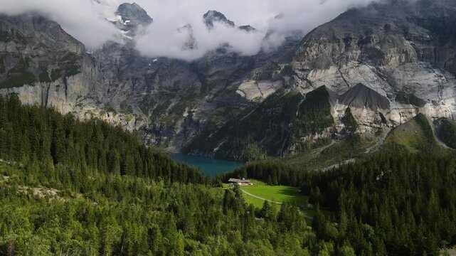 Cinematic aerial view of Oeschinensee, a stunning turquoise alpine lake surrounded by dramatic Swiss Alps, dense forest and green mountain slopes near Kandersteg, Switzerland