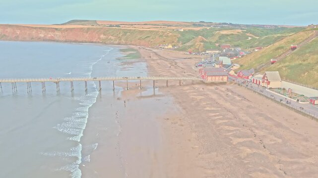 Saltburn Pier extends across the sandy beach beside the Lower Promenade, with the Cliff Lift, Marine Parade and coastal townscape visible at Saltburn-by-the-Sea, England, drone forward flyover.