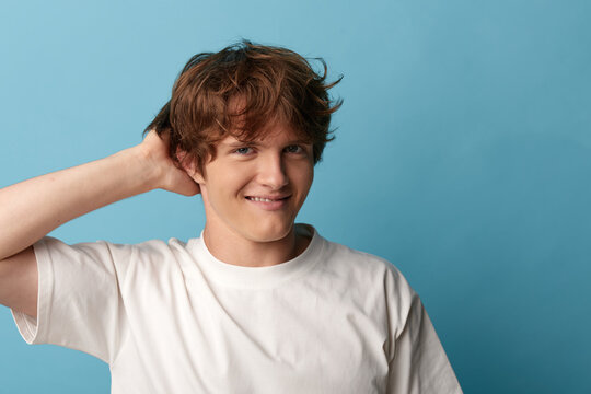 Young man with hand in hair and mischievous smirk against blue background, playful hesitation, casual portrait with confident charm.