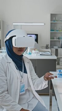 Vertical video of young adult Asian woman scientist wearing VR headset and operating controller in laboratory. Clip fits biotech commercials and modern medical simulation advertising