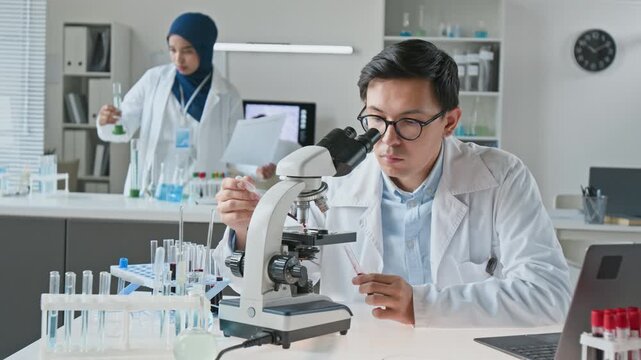Young adult Asian scientist analyzing sample with pipette and looking through microscope in laboratory. Footage fits pharmaceutical research commercials and medical biotech projects