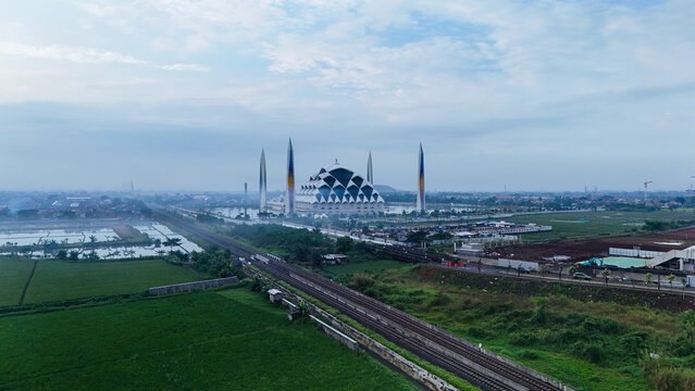 Aerial view of Al Jabbar Grand Mosque surrounded by lush green rice fields, railway tracks, and urban landscape in Bandung, West Java, Indonesia.