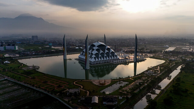 Aerial view of Al Jabbar Grand Mosque surrounded by a reflective pond and lush gardens under a golden sunset sky in Bandung, West Java, Indonesia.