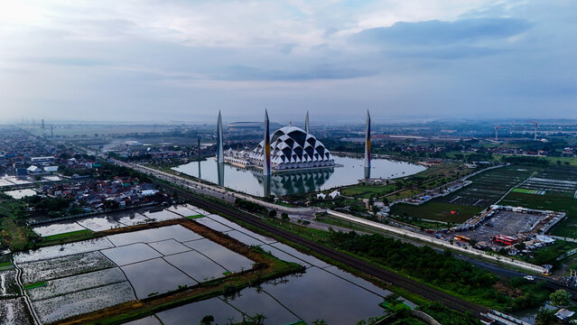 Aerial view of Al Jabbar Grand Mosque surrounded by a reflective pond, lush greenery, and urban landscape in Bandung, West Java, Indonesia.